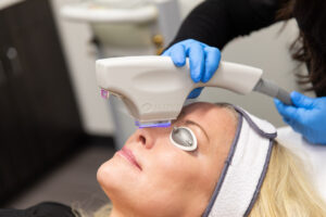 A woman wearing eye protection and receiving treatment on her nose from a professional holding a light therapy tool